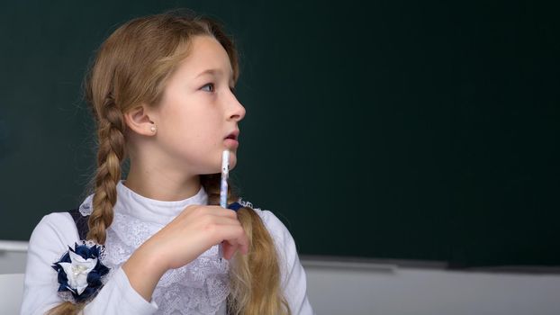 Portrait of pensive schoolgirl. Thoughtful girl holding pen near her mouth looking away. Primary school student posing on background of blackboard. Back to school, education concept