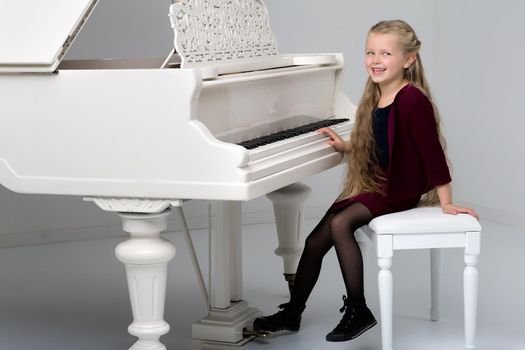 Smiling girl playing grand piano. Full length portrait of adorable long haired little girl sitting at white piano. Cute child having fun with learning to play music instrument