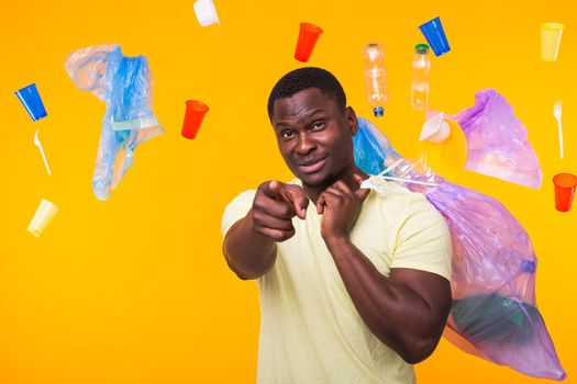 Problem of trash, plastic recycling, pollution and environmental concept - funny african american man carrying garbage for recycling and pointing on you on yellow.