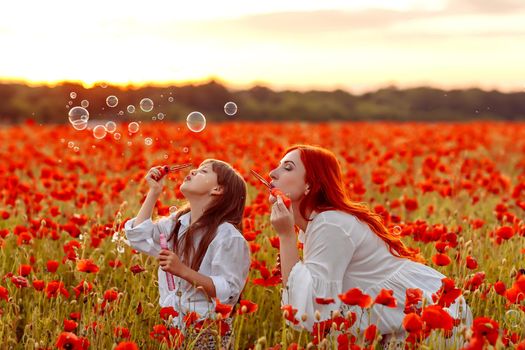 Little happy girl with redhead mother in white dresses blow bubbles on poppy field at warm summer sunset