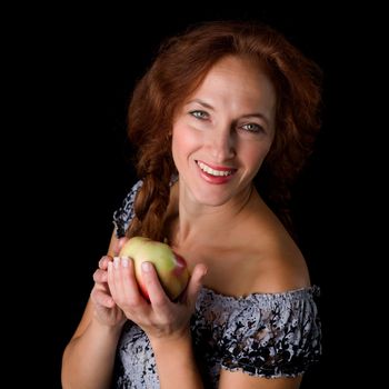 Joyful woman holding fresh apple. Portrait of girl smiling at camera against black background. Happy woman with brown hair posing with ripe healthy fruit in studio