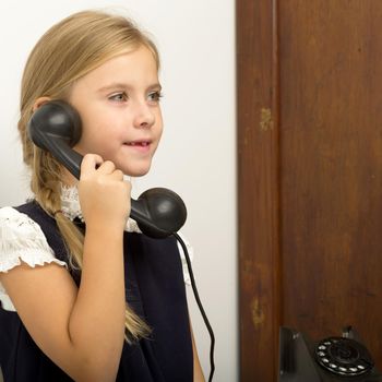 A cute girl is talking on an old phone. A lovely blonde talking on an old-fashioned phone in the studio. Photo of a beautiful child posing in a retro interior
