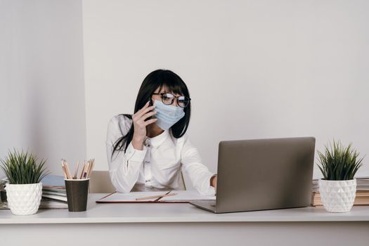 A young woman works remotely in the office with a protective mask during an epidemic. Conducts a video conference and talks on the phone