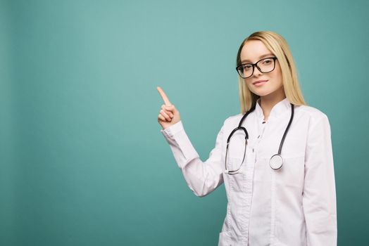 Smiling young medical doctor woman with stethoscope pointing on copyspase. Isolated over blue background.