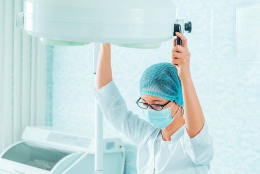 Medical assistant in a mask and cap places a surgical lamp for operation