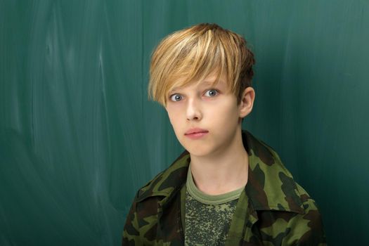 Closeup portrait of confident teenage boy. Blond boy in khaki military clothing. Student wearing camouflage t-shirt and military jacket posing in front of blackboard