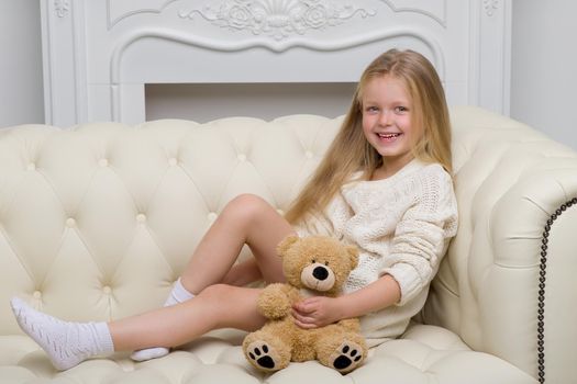 Lovely blonde girl sitting on comfortable sofa hugging teddy bear. Portrait of cute long haired girl wearing warm knitted dress. Adorable happy child resting on couch with favorite stuffed toy