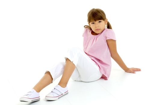 Cute stylish preteen girl sitting on floor. Lovely child in pink t-shirt, white pants and sneakers leaning back on her hands on isolated white background. Brown eyed girl looking seriously at camera