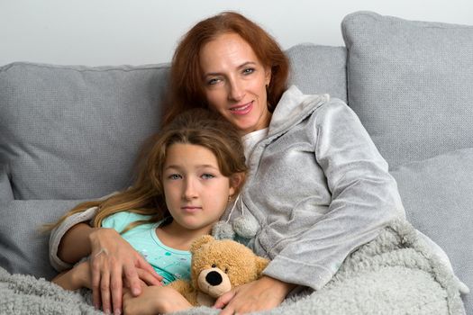 Happy mom sitting on couch with her preteen daughter. Smiling loving mother hugging her daughter lying with teddy bear toy. Woman and child wearing indoor clothes sitting together covered with plaid