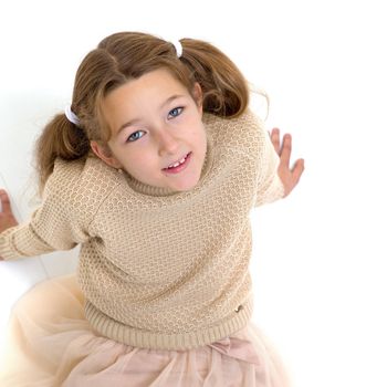 Happy preteen girl sitting on floor leaning back on hands. Beautiful smiling girl with pigtails wearing beige knitted jumper and skirt looking at camera posing against white background.