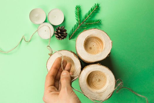 DIY Christmas candle holder made of pine logs, candles, craft rope, fir branches and cones. Hands fasten and tie the rope. Step-by-step instructions flat lay, step 3.