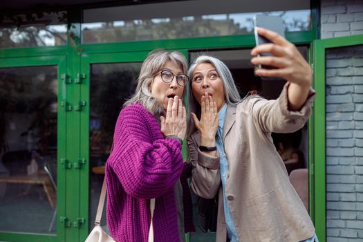 Funny senior ladies friends take selfie covering mouthes with palms with mobile phone on modern city street. Long-time friendship relationship