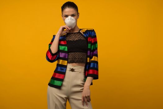 Beautiful slim girl fashion model posing in a protective respirator on a yellow background. LGBT community rainbow color jacket