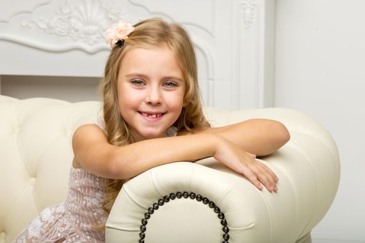 Beautiful happy girl sitting on sofa. Retro style closeup portrait of smiling blonde girl in nice dress sitting on comfortable couch. Adorable happy seven years old kid posing in studio