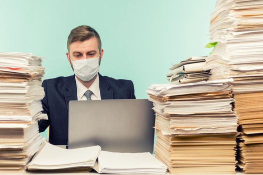 A male accountant or company manager works in an office in a pandemic in view of the accumulated paper work. A protective medical mask is on the face. On the desktop are large stacks of documents. - image