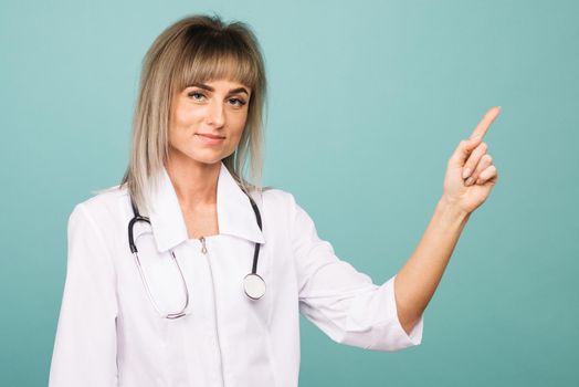 Smiling young female doctor with a stethoscope points up her fingers on a blue background