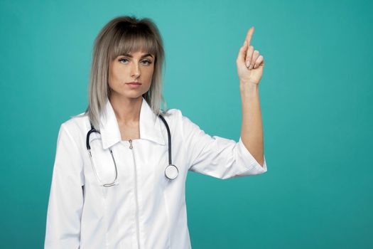 Smiling young female doctor with a stethoscope points up her fingers on a blue background