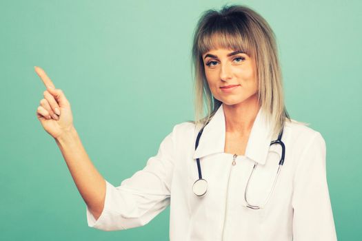 Smiling young female doctor with a stethoscope points up her fingers on a blue background