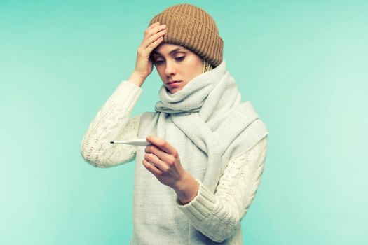 The girl has symptoms of coronavirus. Young woman having flue taking thermometer against blue background. Beautiful lady is sick with a high temperature and headache. Cold, flu concept.