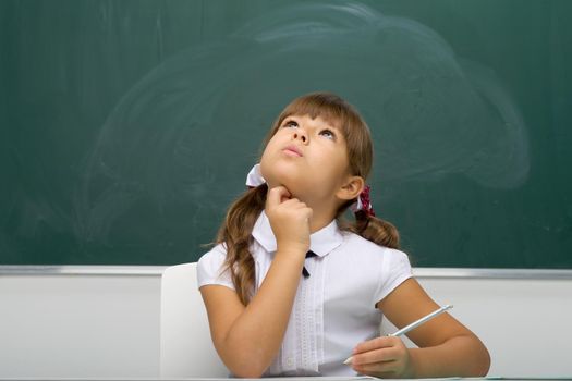 A pensive girl is sitting at the table, looking up. Elementary school student beautiful girl with ponytails posing against the background of the blackboard in the classroom. School and education concept