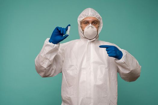 A male doctor in protective overalls and a mask pointing at ampoule with a vaccine in his hands. Coronovirus protection concept in China. - image