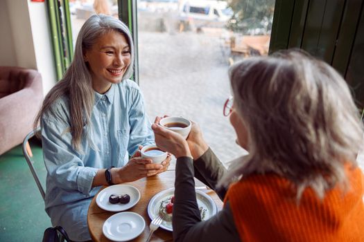 Pretty happy mature women companions with natural grey hair hold cups of hot drinks sitting at small table in cozy cafe. Long-time friendship