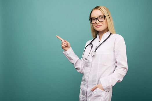 Smiling young medical doctor woman with stethoscope pointing on copyspase. Isolated over blue background.