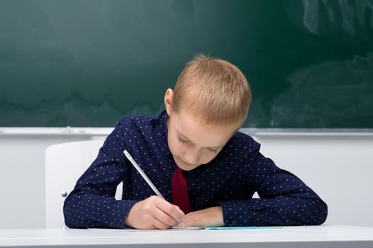 Schoolboy writing in notebook. School student in blue shirt and tie sitting at desk on background of blackboard in classroom. Back to school, education concept