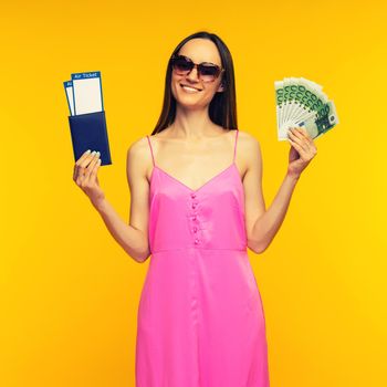 Slender girl in a pink dress and sunglasses holding passport with air ticket and hundred euro bills on a yellow background