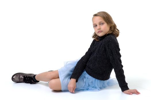 A beautiful girl in a black jumper and a blue fluffy skirt sits on the floor. Pretty blonde woman in fashionable clothes posing in studio on white background