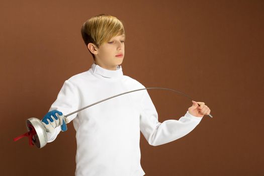 Boy fencer in white fencing costume with rapier. Closeup portrait of teenage boy wearing white fencer uniform posing against brown background. Teenager practicing in fencing