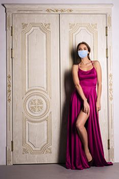 Fashion portrait of a woman posing in protection mask and beautiful evening dress, near the big white doors. Luxurious interior, perfect figure and hair girl