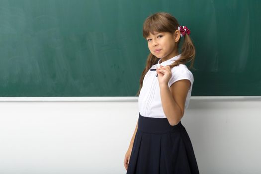 Pretty girl posing at blackboard. Portrait of beautiful schoolgirl in school uniform standing at green chalkboard with piece of chalk in her hand and looking at camera. School and education concept