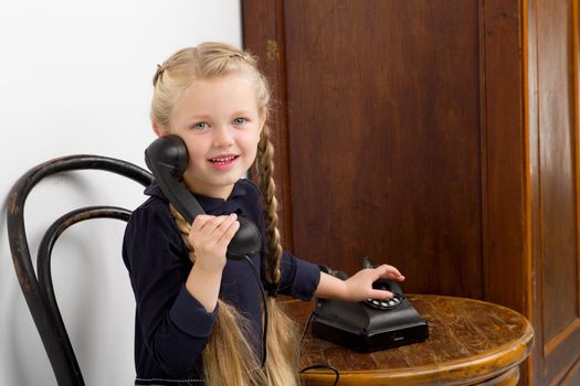 Blonde girl talking by old black phone. Lovely girl with braids sitting on chair in vintage room interior with old wooden furniture. Cute six years old kid speaking on retro telephone