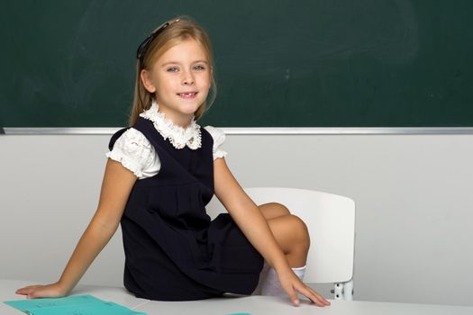 Adorable schoolgirl sitting on desk in classroom. Smiling blonde girl student in school uniform sitting in front of blackboard. School and education concept