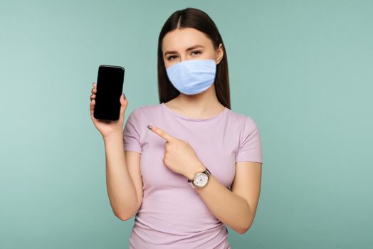 Young sad girl in mask points at smartphone on blue background. The concept of Internet dependence on social networks. Coronavirus Outbreak Defense Concept