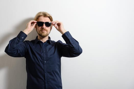 Stylish man in a blue shirt and sunglasses standing near a white wall and a hand holding glasses