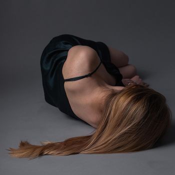 thin caucasian girl with long brown hair in dress lies on gray floor with her head turned away. Depression, anxiety, solitude concept of lonely woman. female wearing dark green dress lying on ground