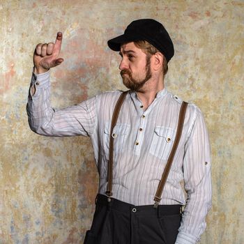 Portrait of handsome young man in white shirt and bow tie adjusting his suspenders and smiling at camera while standing isolated near old wall