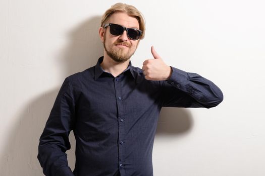 Attractive man in black sunglasses standing near a white wall holding up thumbs up.