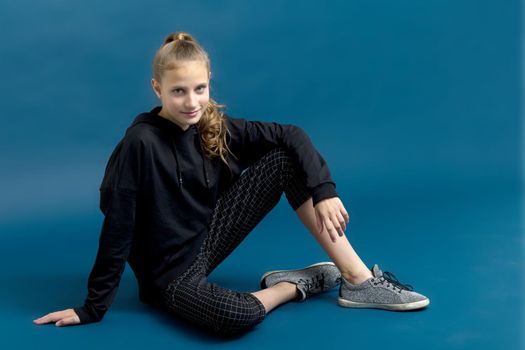 Teen girl in black hoodie sitting on floor. Portrait of beautiful serious teenager dressed black sweatshirt, breeches and sneakers posing against blue background in studio