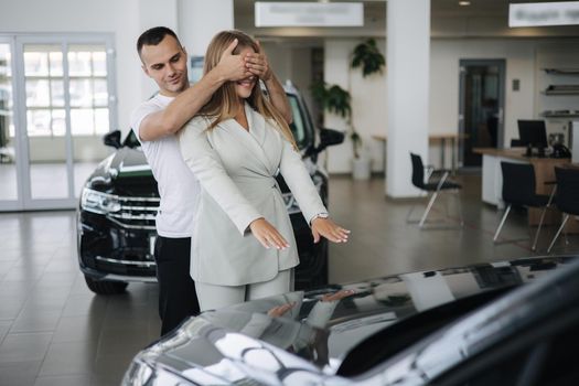Man close eyes to his wife and makes a surprise buying car. Man and woman in car showroom.