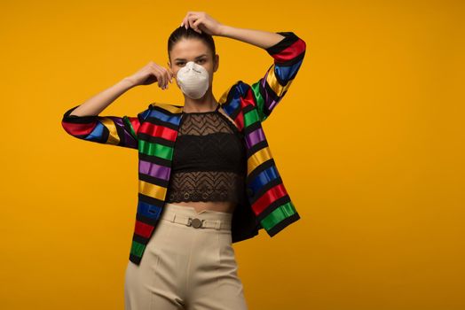 Beautiful slim girl fashion model posing in a protective respirator on a yellow background. LGBT community rainbow color jacket