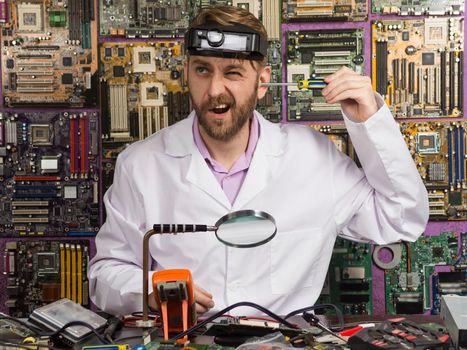 Funny engineer in white coat holding a screwdriver in his ear while sitting at the table in his laboratory.