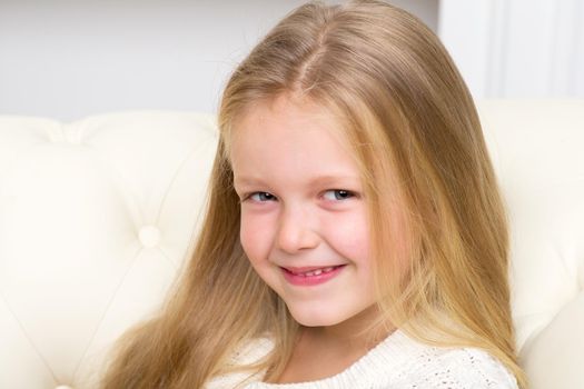 Close up portrait of lovely blonde girl sitting on comfortable sofa. Cute smiling long haired little girl having fun at home. Adorable happy child resting on couch