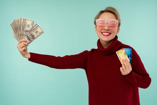 Portrait of happy young woman in pink sunglasses holding credit card and money in hand smiling and looking at camera on isolated blue background feeling positive and enjoy - image
