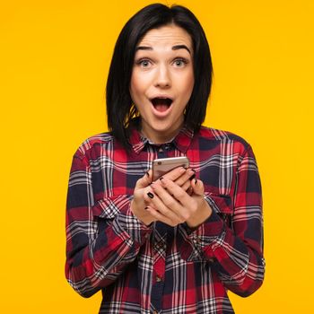 Excited laughing woman in plaid shirt standing and using mobile phone over yellow background - Image