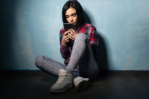 Young sad girl suffering from dependence on social networks sitting on the floor with a smartphone