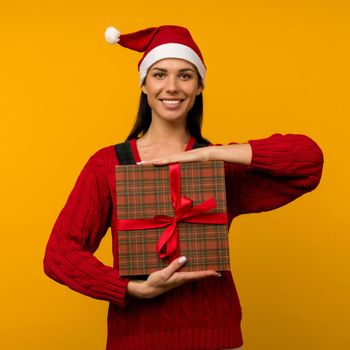 Happy excited young woman in santa claus hat with gift box over yellow background - image