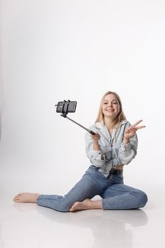 happy female making self portrait with smartphone attached to selfie stick. young pretty blond girl with long hair smiling, using cell phone isolated on white background, showing peace sign gesture